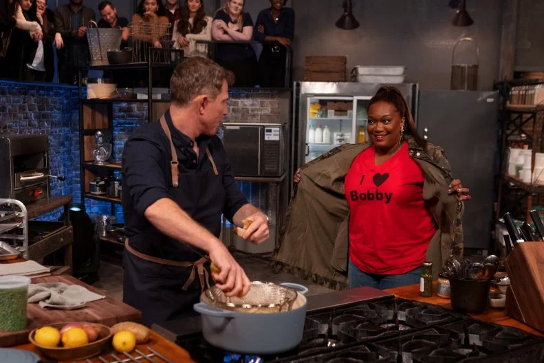 Co-host Sunny Anderson shows Bobby Flay an “I Love Bobby” t-shirt beneath her jacket as he cooks during round two, as seen on Beat Bobby Flay, Season 30.