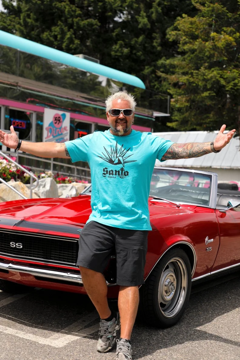 Host Guy Fieri posing in front of his Camaro outside of 59'er Diner in Leavenworth, Washington, as seen on Diners, Drive-ins, and Dives, season 34.