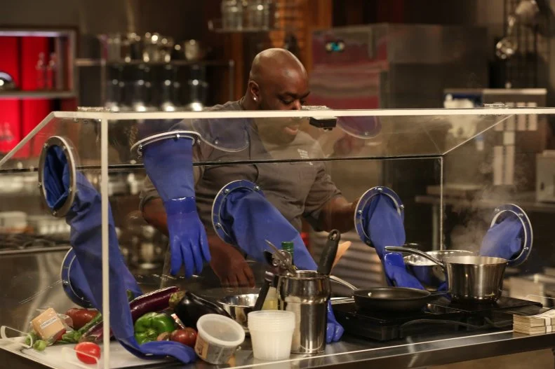 Chef Kenny Gilbert works with a sabotage that forces him to do all of his cooking in a plexi box station, as seen on Food Network's Cutthroat Kitchen, Season 9.
