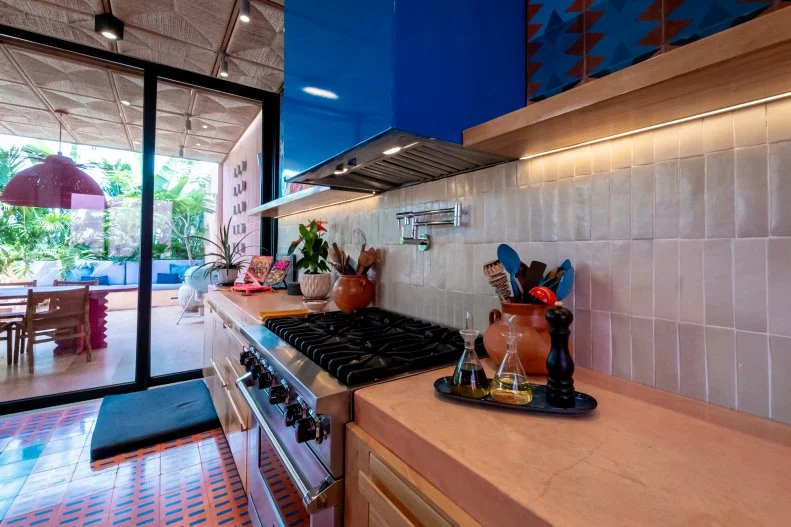 The metal framed glass doors can be opened to immediately create one large room blurring the kitchen and an outdoor dining space. Bold shades of wine and cobalt blue coexist with plenty of wood, clay and other nature fibers like the palm woven tiles above.