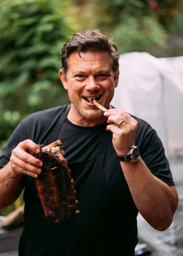 Tyler Florence bites into a rack of grilled ribs outside.