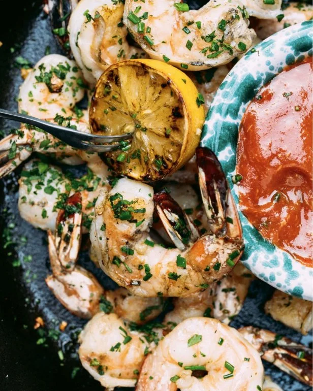 A platter of Tyler Florence's grilled shrimp cocktail with grilled lemons, plated with a blue and white bowl of cocktail sauce.