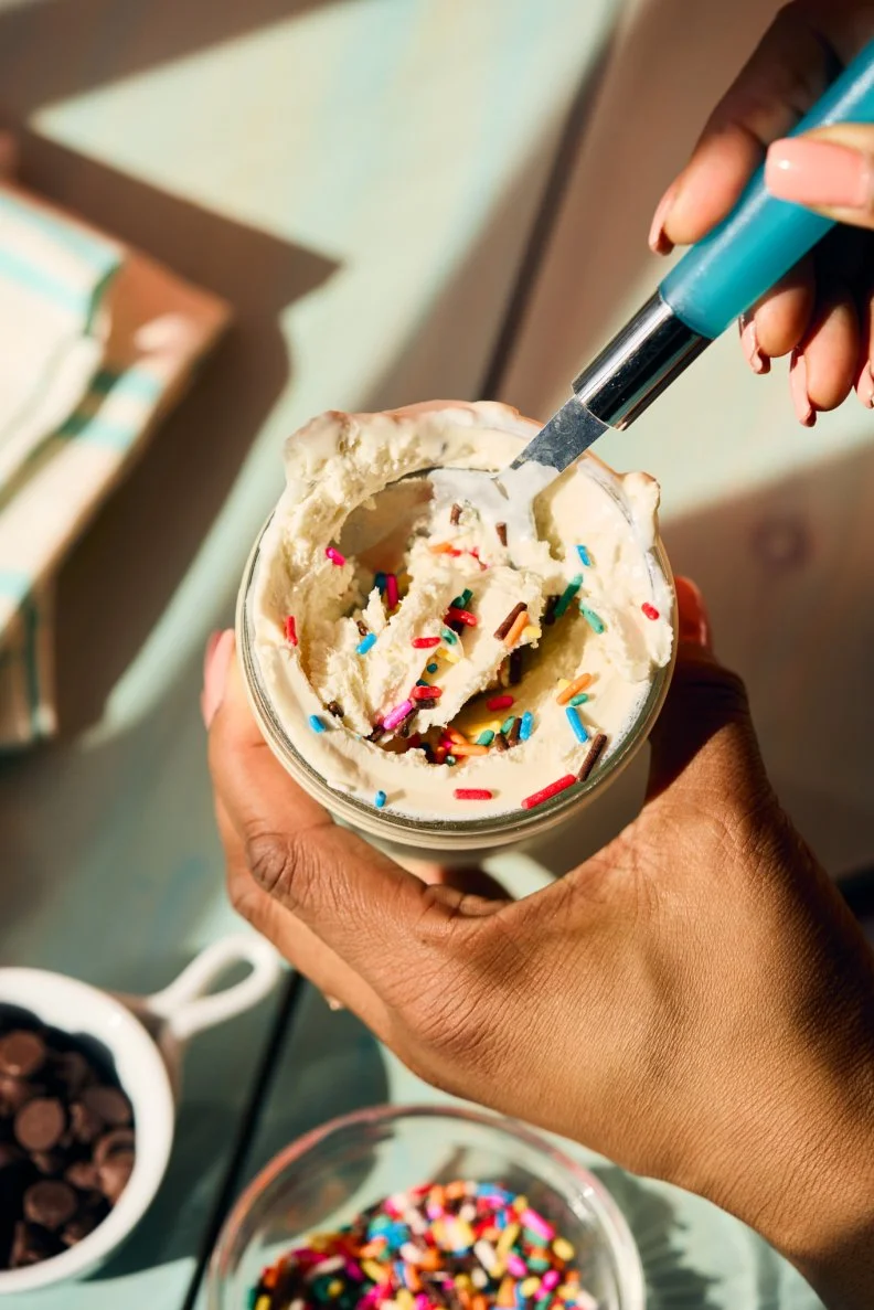 A close-up of somebody using a spoon to dip into a small jar of vanilla ice cream with sprinkles 