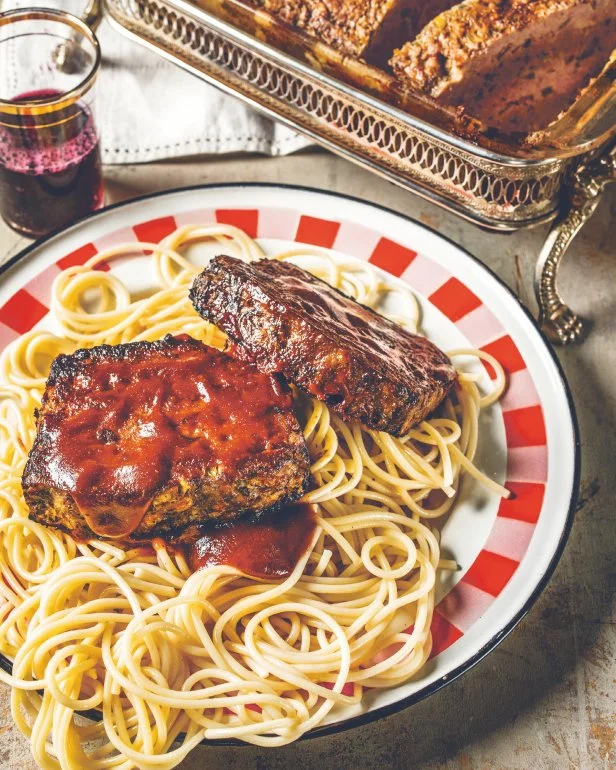 Meatloaf on top of spaghetti, plated on a white-and-red checkered plate next to a metal serving dish.