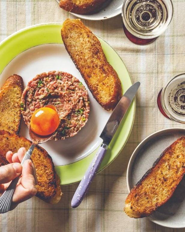 A hand holding a spoon drops a hot-sauce cured egg yolk onto a neat bison tartare plated with toasted bread on a green plate.