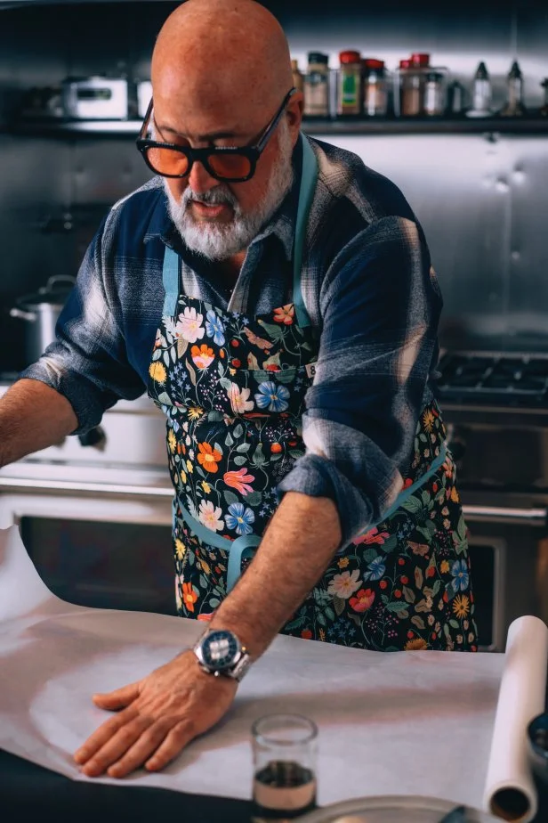 A bald, bearded man in a floral apron smoothes parchment paper.