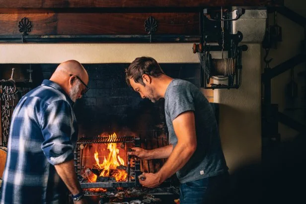 Two men standing at a traditional kitchen hearth with an open fire.