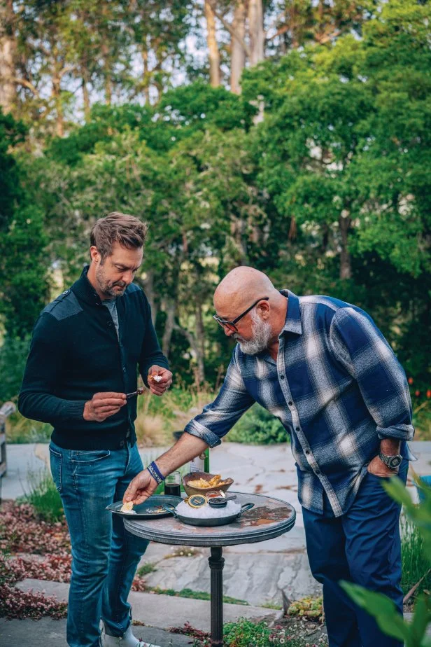 Two men standing at a table with caviar on ice and a bowl of chips.