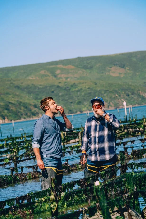 Two men in waders eating oysters while standing in an oyster bed.