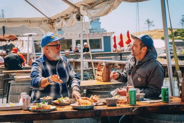 Two men seated at a wharfside table covered with plates of seafood.