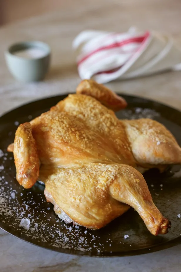 A crispy, skin-on roast chicken on a serving platter with a small bowl of salt and a dishtowel in the background.