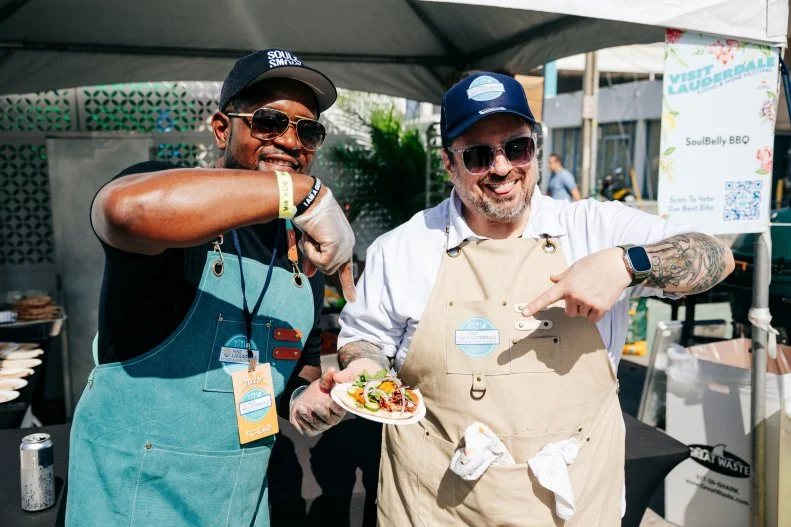 Chefs D'Andre Carter and Bruce Kalman pose and point to a dish outside the Soul Belly tasting tent at the Visit Lauderdale Food & Wine Festival.