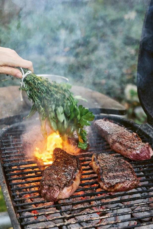 A hand uses an herb bundle to brush a charred steak over a small coal grill. 