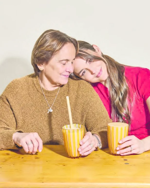 Chef Mika Leon (left) and her mother pose (right) for a photo while drinking mango smoothies out of glass cups. Mika leans on her mother's shoulder.