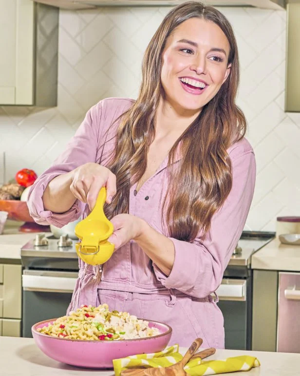 Chef Mika Leon, donned in a pink jumpsuit, uses a yellow citrus press to squeeze juice into a pink bowl of macaroni salad.