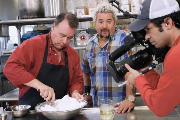 Guy Fieri looking in to the camera as Chef/Owner Rusty Winkstern whisking egg whites for the waffle batter during Guy's original DDD visit to Monument Cafe in Austin, Texas, as seen on Food Network's Triple D Nation, season 8.