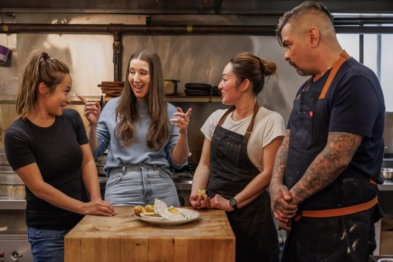 Jen Agg and Aaron Sanchez smiling with Joanna L and Jeanette L in the kitchen with Chili Crisp Crab Spring Rolls dish on the table in front of them