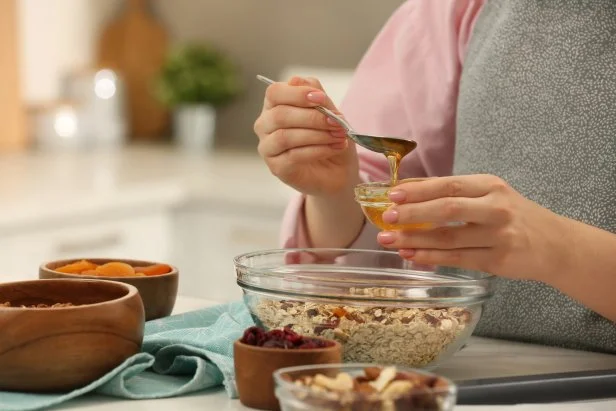 Making granola. Woman adding honey into bowl with mixture of oat flakes and other ingredients at table in kitchen, closeup