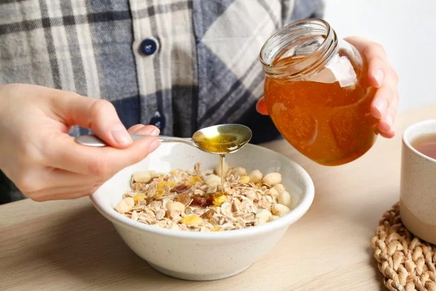 Woman putting honey into bowl with muesli at wooden table, closeup