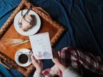 View from above of a mother holding a handmade Mother's Day card with a tasty breakfast in bed. She is on a blue colored sheet.