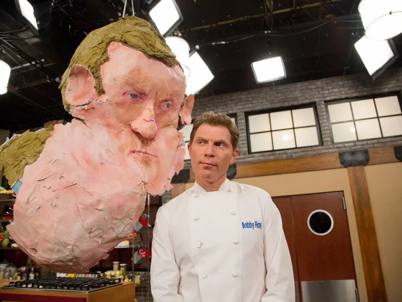 Blue Team Leader Chef Bobby Flay posing with the giant pinata replica of his head, during the Surprise Party Challenge. Each team member must guess the flavor of the candy they pick up from the pinatas they burst, and infuse the dishes the chefs demonstrate for them with the individual flavor each gets, as seen on Food Network's Worst Cooks in America, Season 5.