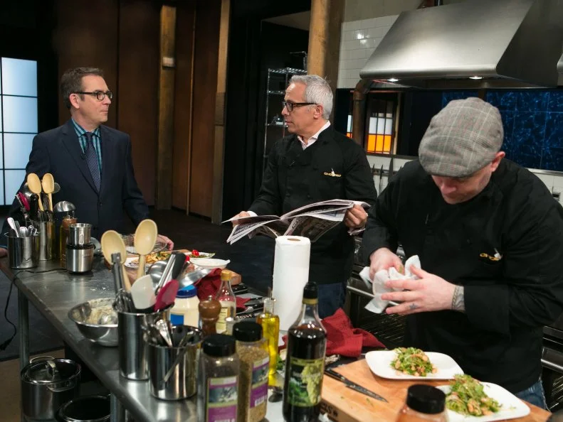 Chef Geoffrey Zakarian reads the newspaper while working on his dish that must include: eel, pepihuates, shaved coconut and sea beans, as seen on Food Network's Chopped, Season 21.