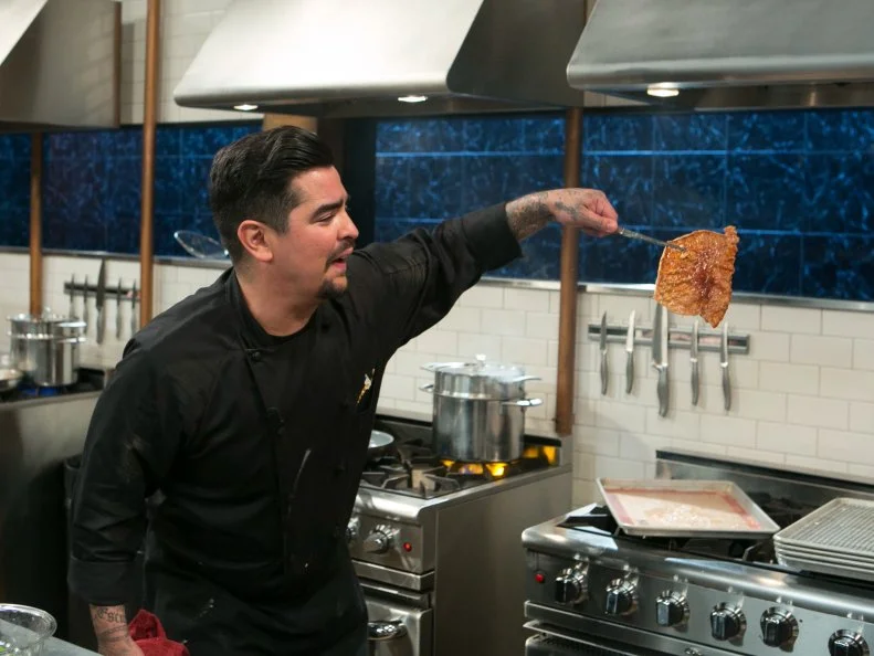 Chef Aaron Sanchez shows his crispy pork skin off to the cameras as he works on his dish that must include: whole suckling pig, kebab sauces, fiddlehead ferns and fresh corn tortillas, as seen on Food Network's Chopped After Hours, Season 21.