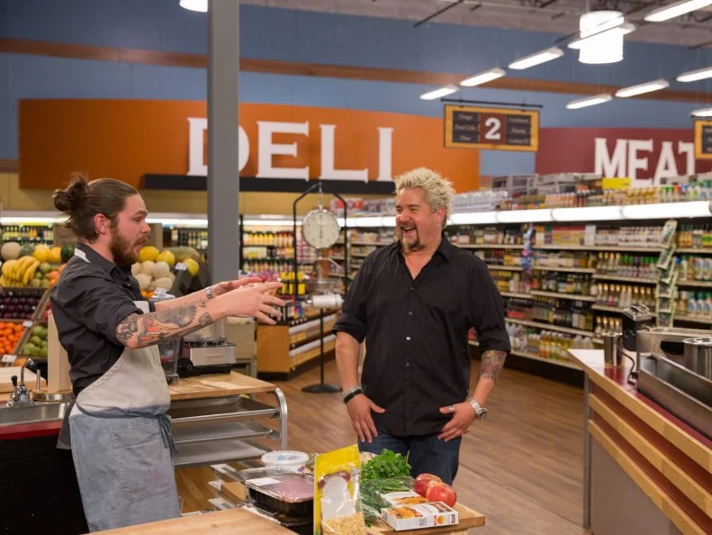Host Guy Fieri talks with Chef Phillip Franklin Lee as he prepares his dish during Game 3, Grocery Bowl, as seen on Food Network's Guy's Grocery Games, Season 7.