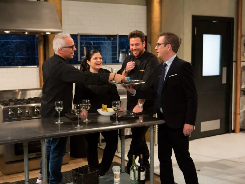 Chopped host Ted Allen theatrically delves chef Geoffrey Zakarians machiatto while Alex Guarnaschelli and Scott Conant  laugh, as seen on Food Networks Chopped, season 27 webisode