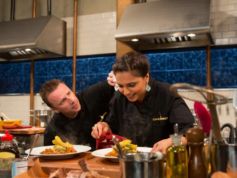 Chef Marc Murphy dabs sweat away from chef Maneet Chauhans forhead  as she works on her dish that must include: North Carolina style bbq sauce, pork spare ribs, rainbow chard and avacados , as seen on Food Network's Chopped After Hours, Season 23.