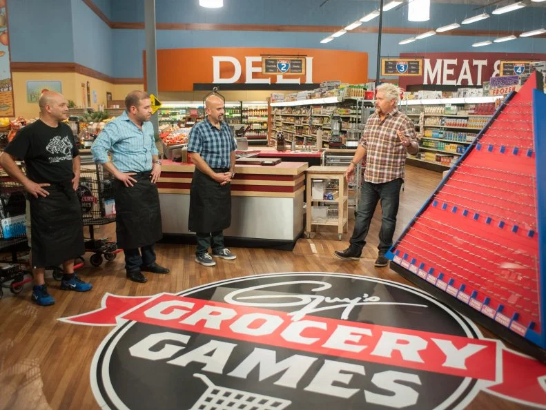 Host Guy Fieri explains the "Food Pyramid" game to contestants George Prisco, Aristo Boutsikakis and Jay Hajj, as seen on Food Network's Guy's Grocery Games, Season 8.