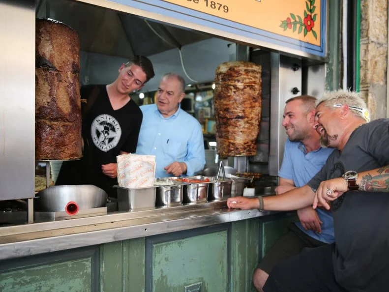 Guy Fieri, Aristo, chef/owner Shiro Bairaktaris of Bairaktaris Tavern in Athens, Greece watch as Hunter Fieri slices the meat for a Gyro as seen in Food Network's Guy's European Vacation episode 1.