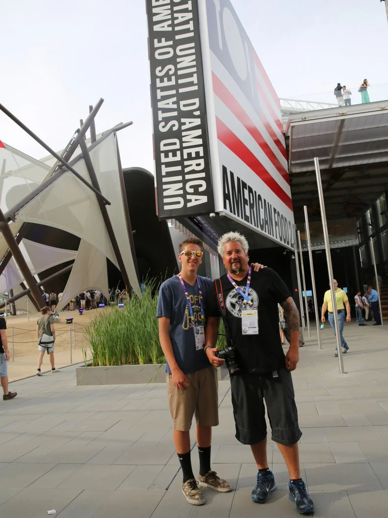 Portrait shot of Guy and Hunter Fieri standing next to the American Food sign at the Food World's Fair Expo in Milan, Italy as seen on Food Network's Guy's European Vacation episode 3.