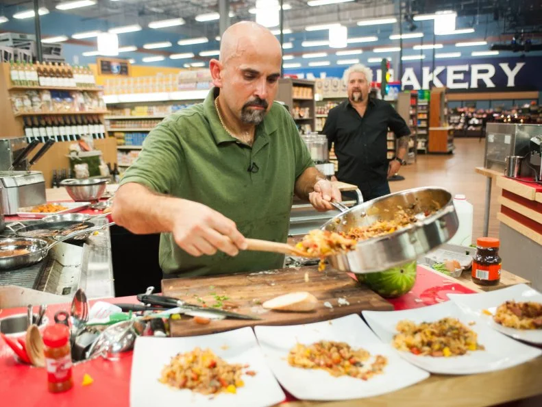 Contestant Jay Hajj finishes preparing his dish as host Guy Fieri looks on, as seen on Food Network's Guy's Grocery Games, Season 8.