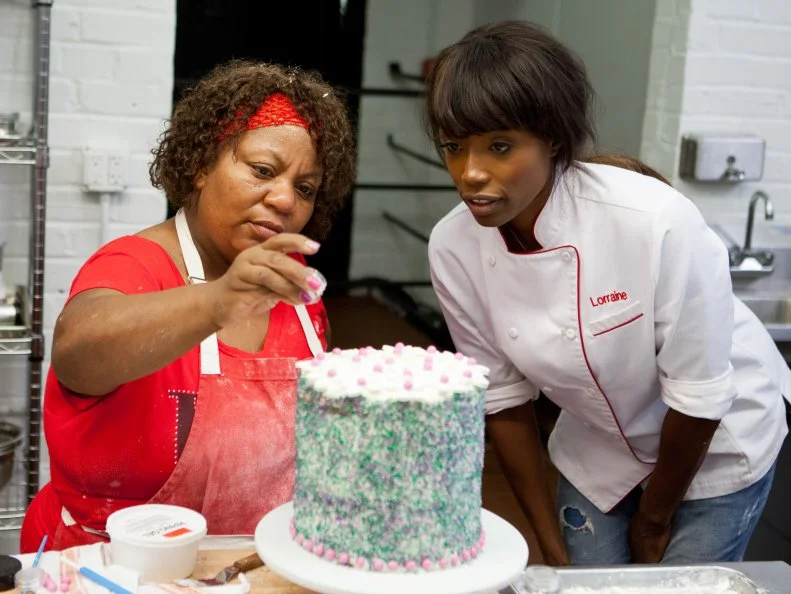 Contestant Carla Johnson works on final touches of her cake while Host Lorraine Pascale watches and offers help, as seen on Food Network's Worst Bakers In America, Season 1.