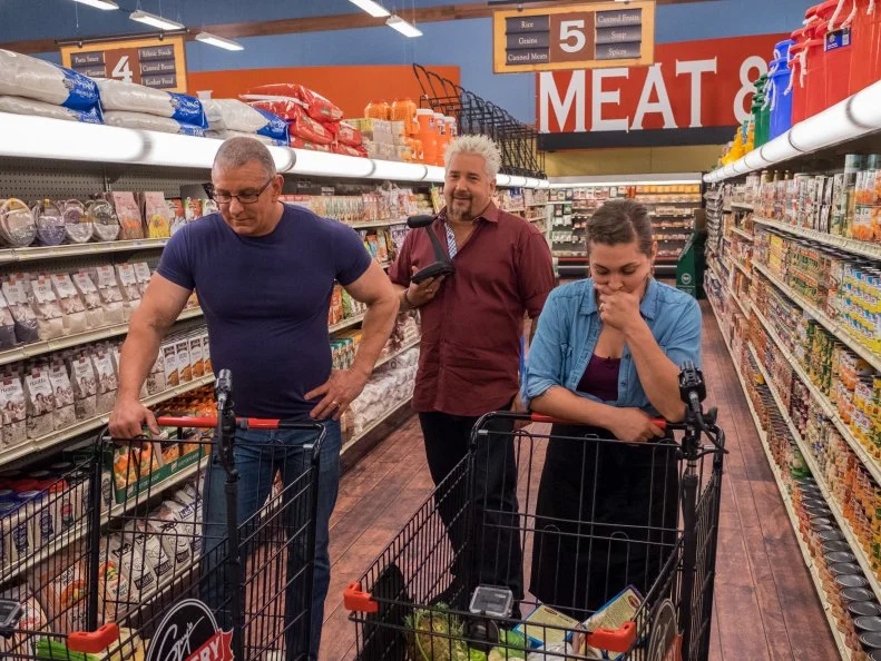 Contestant Samantha Mitchel and judge Robert Irvine look inside their grocery carts as host Guy Fieri speaks into the PA system, as seen on Food Network's Guy's Grocery Games, Season 11.