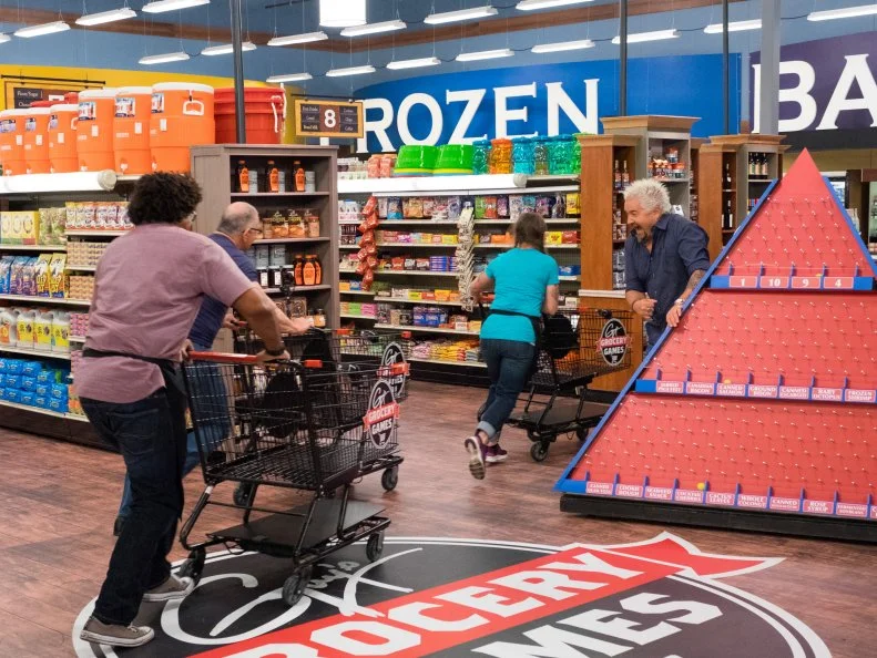 Contestants Samantha Mitchel, Stephen Jones and judge Robert Irvine race past host Guy Fieri, as seen on Food Network's Guy's Grocery Games, Season 11.