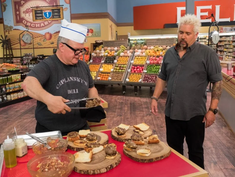 Contestant Zane Caplansky finishes adding his burgers onto toasted buns as host Guy Fieri looks on, as seen on Food Network's Guy's Grocery Games, Season 12.