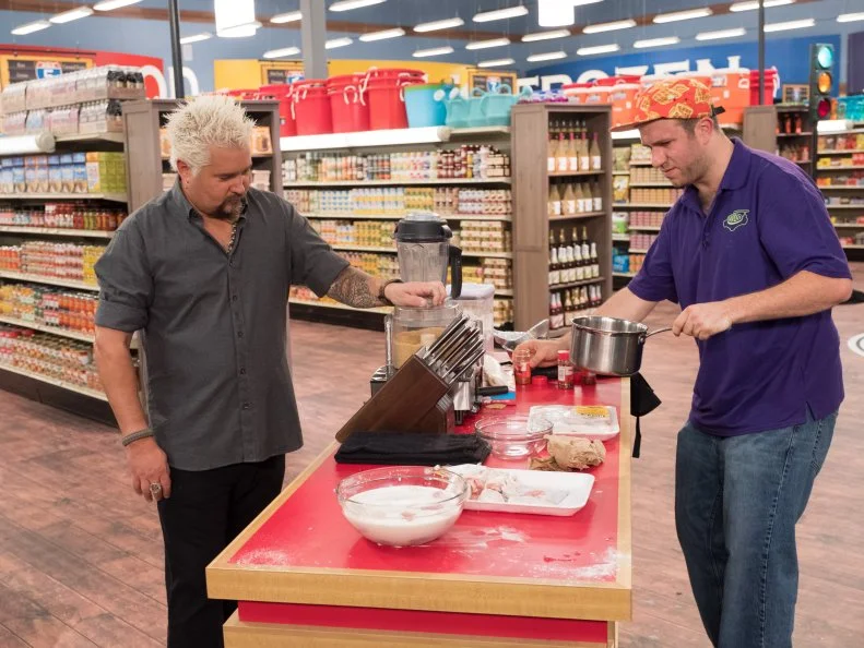 Host Guy Fieri looks on as contestant Adam Gold prepares his dish, as seen on Food Network's Guy's Grocery Games, Season 12.