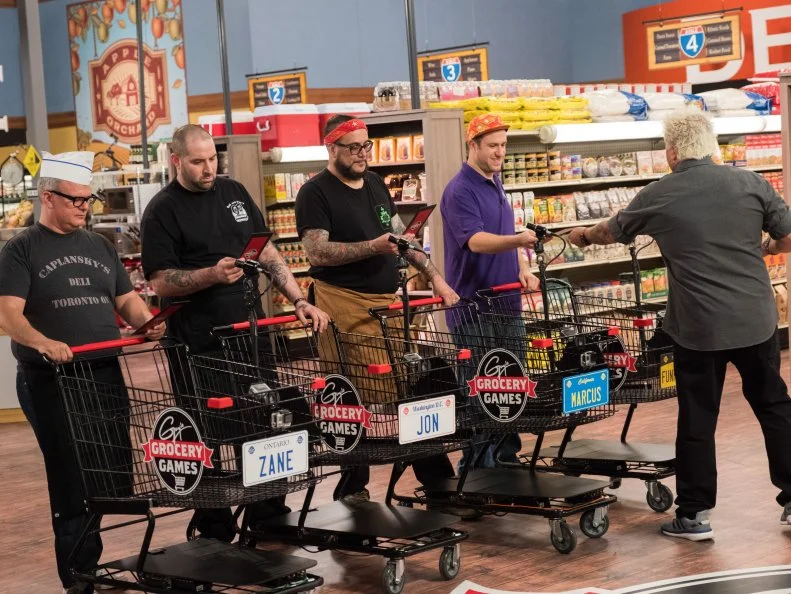 Host Guy Fieri hands out the lists  to contestants Zane Caplansky, Jon Taub, Marcus Christiana-Beniger and Adam Gold for the Flip This LIst game, as seen on Food Network's Guy's Grocery Games, Season 12.