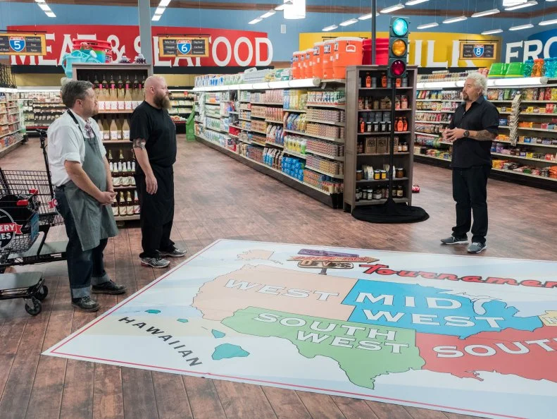 Contestants Peter Campbell and Louie Finnan listen to instructions from host Guy Fieri for the "Regional Road Trip" game, as seen on Food Network's Guy's Grocery Games, Season 12.