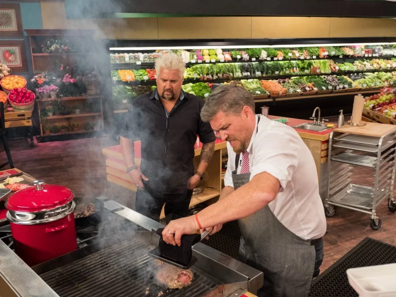 Host Guy Fieri checks on contestant Peter Campbell as he has trouble grilling his burgers, as seen on Food Network's Guy's Grocery Games, Season 12.