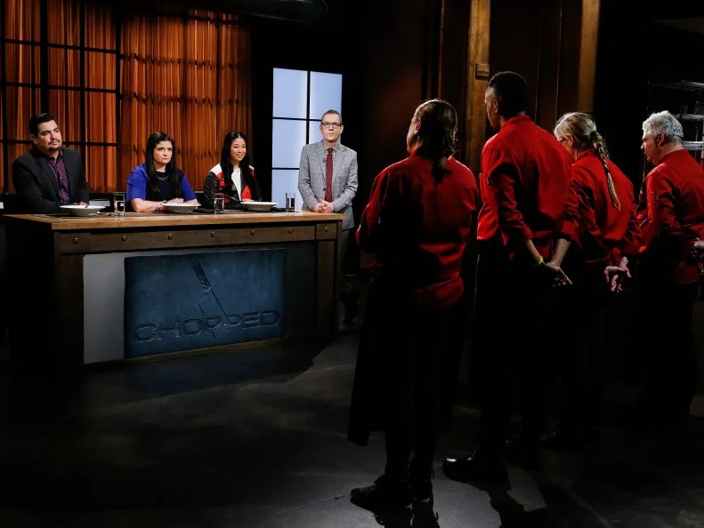 From left, judges Aaron Sanchez, Alex Guarnaschelli and Angie Mar and host Ted Allen face chefs Silvia Baldini, Andre Fowles, Jackie Sappington and Adam Sappington during the appetizer round tasting, as seen on Food Network's Chopped, Season 28.