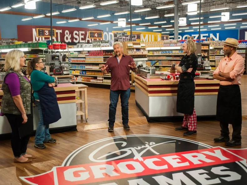 Host Guy Fieri explains the game "Express Lane" to contestants (L-R) Lee Ann Miller, Jodie Rubin, Chase Meneely and Eskender Aseged, as seen on Food Network's Guy's Grocery Games, Season 9.