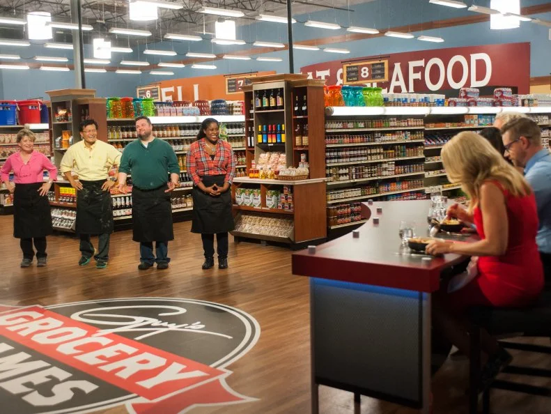 Contestants Mary Schiller, Tom Lin, Robbie Jester and Jamie Gadson wait to hear from host Guy Fieri and judges Brandi Milloy, Richard Blais and Melissa D'Arabian as they sample contestant Jamie Gadson's chicken stir fry, as seen on Food Network's Guy's Grocery Games, Season 9.