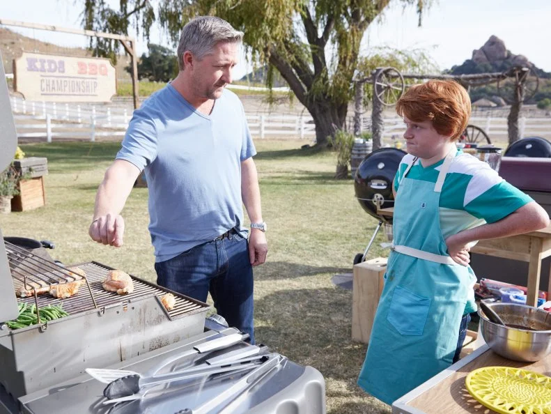 Guest Judge Tim Love with contestant Jacob Scott, as he prepares his dish for the Chicken BBQ Challenge, as seen on Food Network’s Kids BBQ Challenge Season 1.