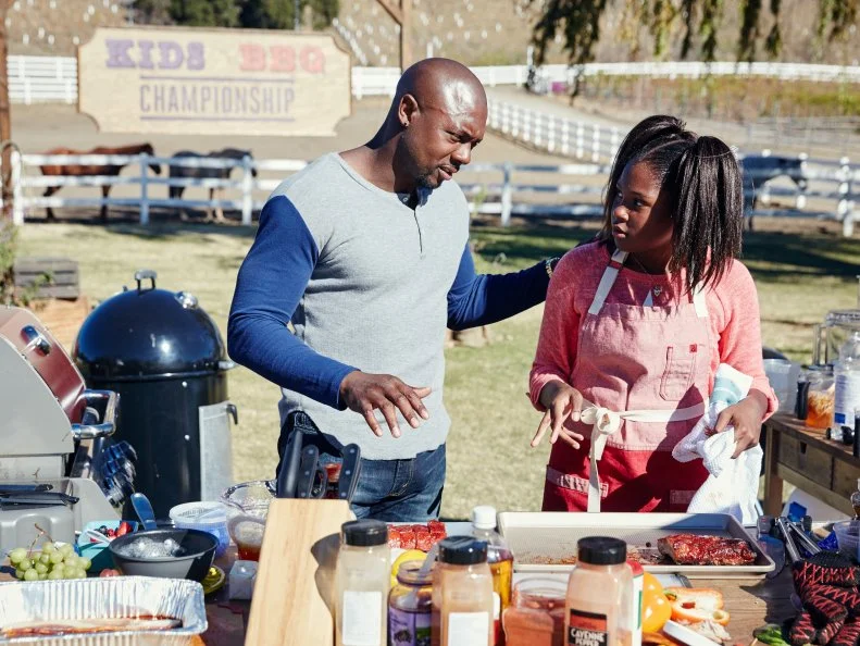 Host Eddie Jackson speaks with Contestant Paris Hale as she prepares her dish for the Pit Master BBQ Challenge, as seen on Food Network’s Kids BBQ Challenge Season 1.