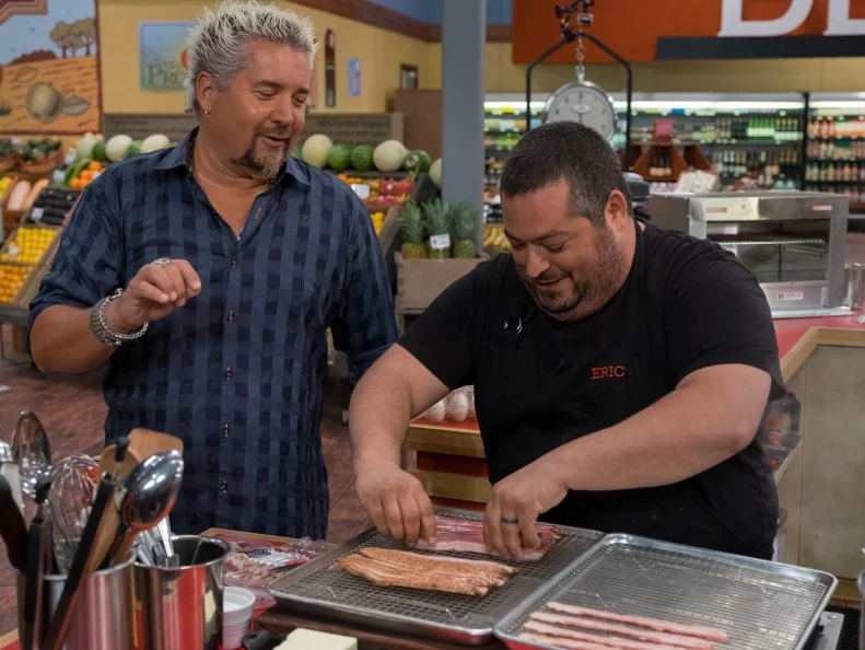 Host Guy Fieri checks on contestant Eric Greenspan as he lays out bacon, as seen on Food Network's Guy's Grocery Games, Season 10.