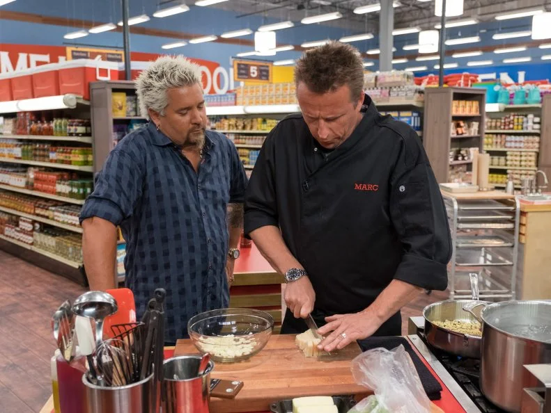 Host Guy Fieri checks on contestant Marc Murphy as he cuts cheese, as seen on Food Network's Guy's Grocery Games, Season 10.