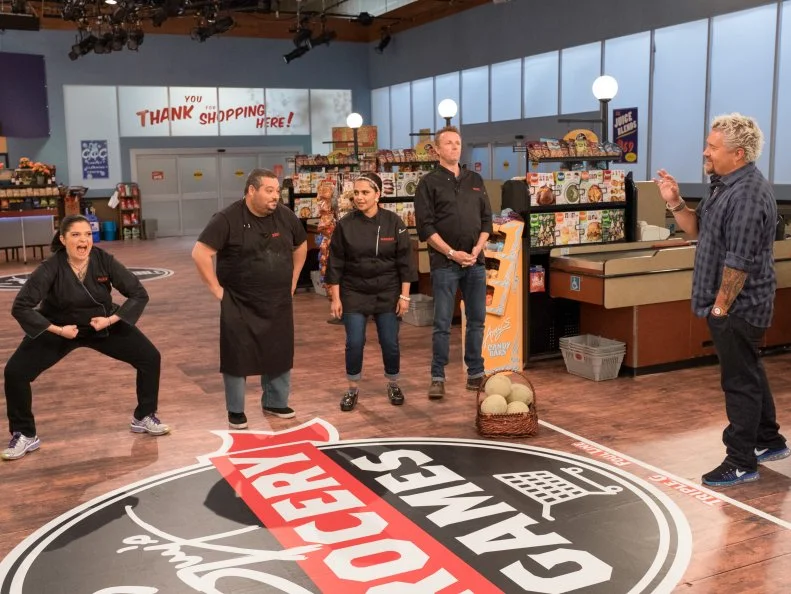 Contestant Alex Guarnaschelli flexes as Eric Greenspan, Maneet Chauhan and Marc Murphy listen to host Guy Fieri explain the "Gorcery Bowl" game, as seen on Food Network's Guy's Grocery Games, Season 10.
